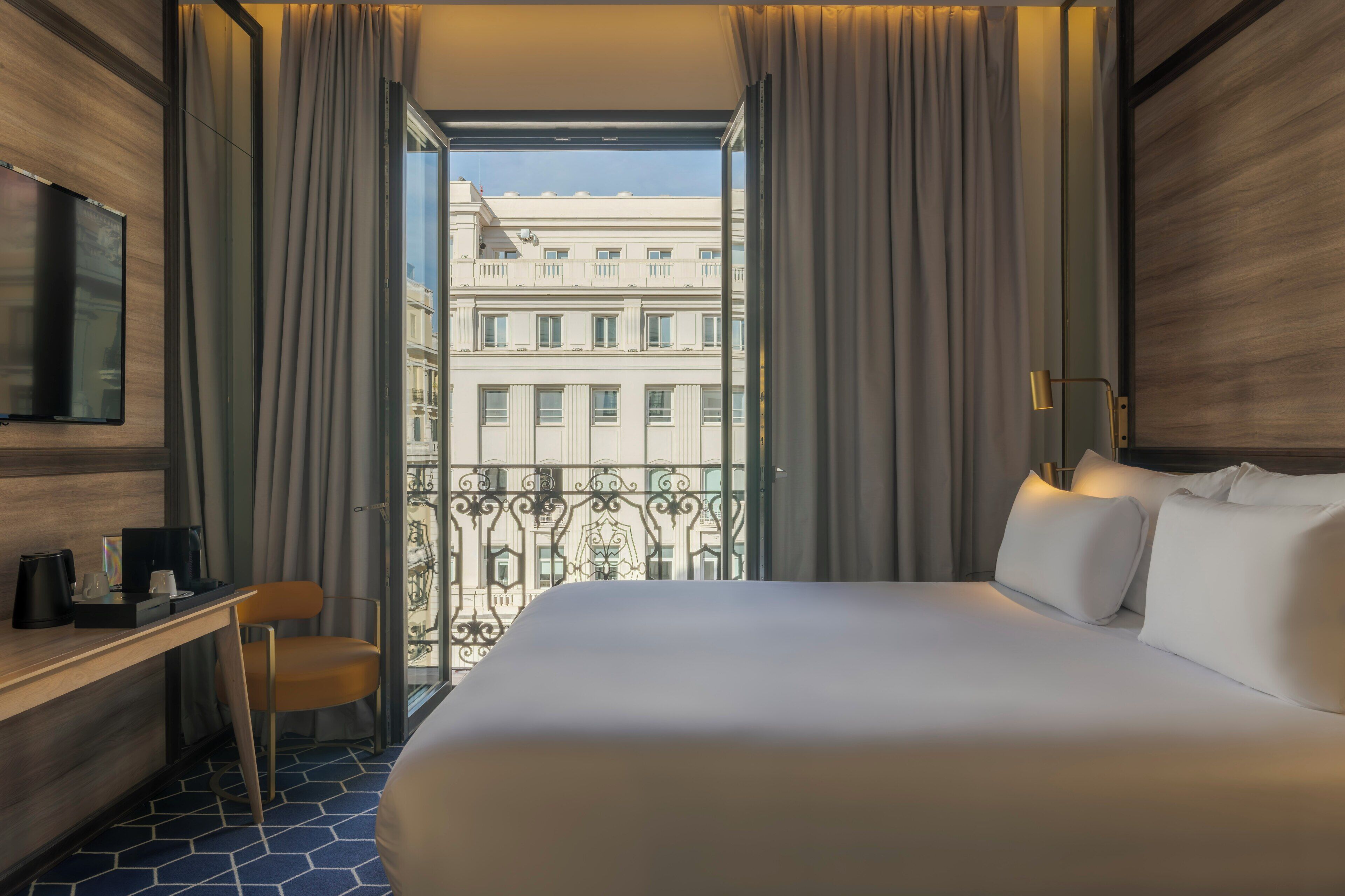 A hotel room with a bed, a desk, a chair, and a view of a building through a window.