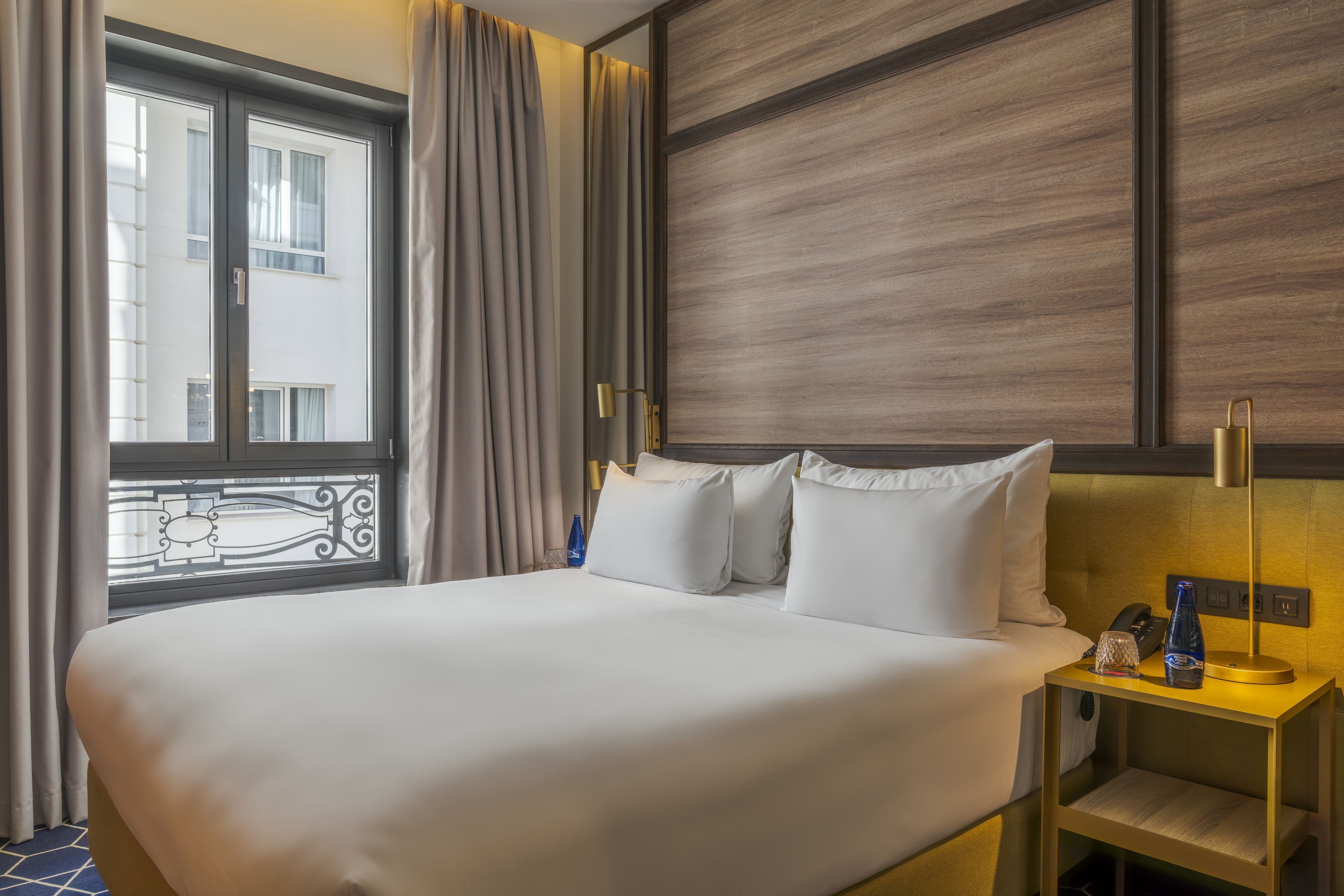 Una habitación de hotel con una cama grande, una mesita de noche con lámpara, una botella y un vaso.