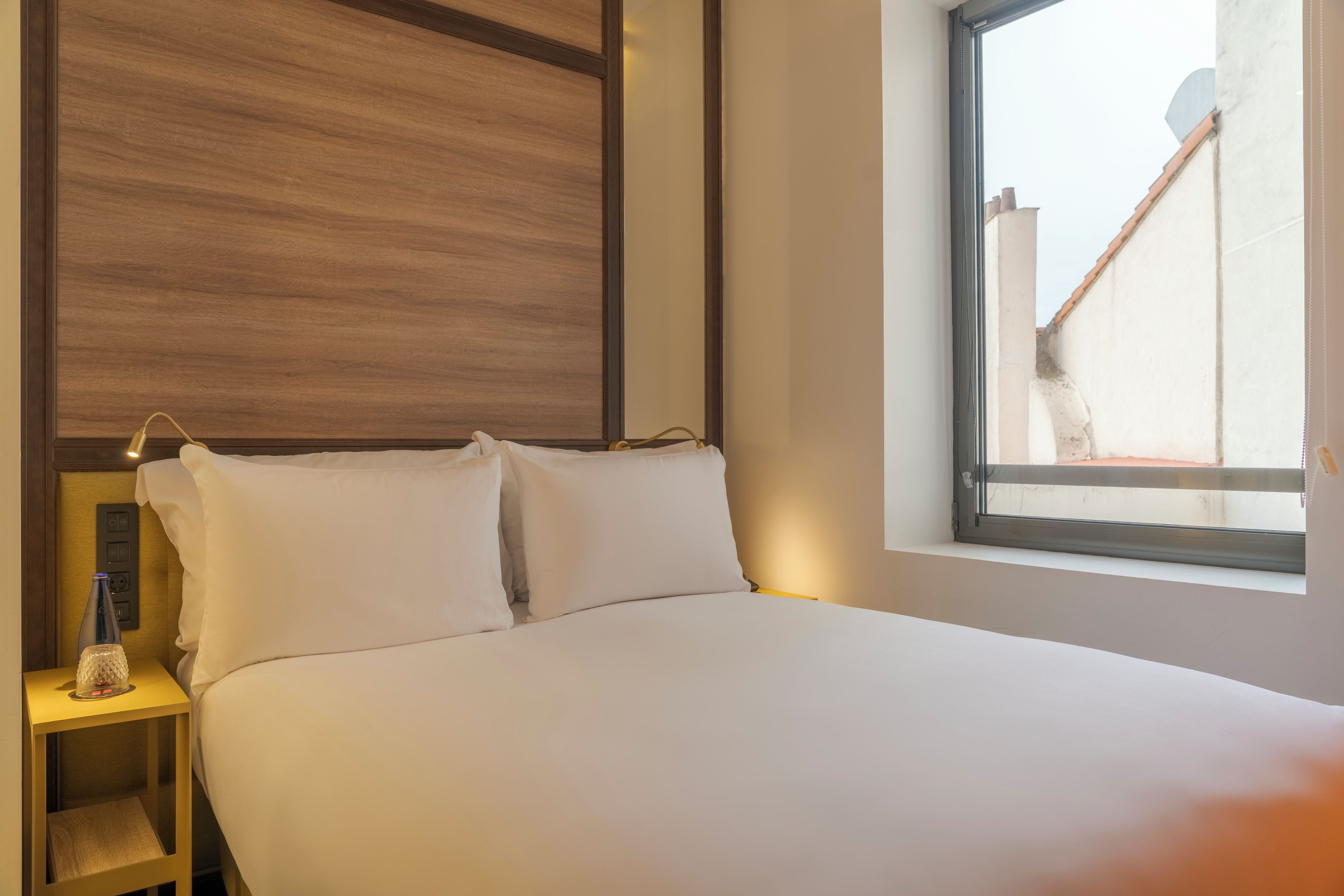 A hotel room with a bed, a nightstand, a bottle, and a window with a view of buildings.