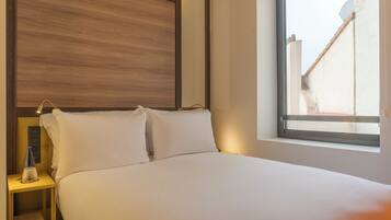 A hotel room with a bed, a nightstand, a bottle, and a window with a view of buildings.