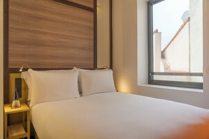 A hotel room with a bed, a nightstand, a bottle, and a window with a view of buildings.