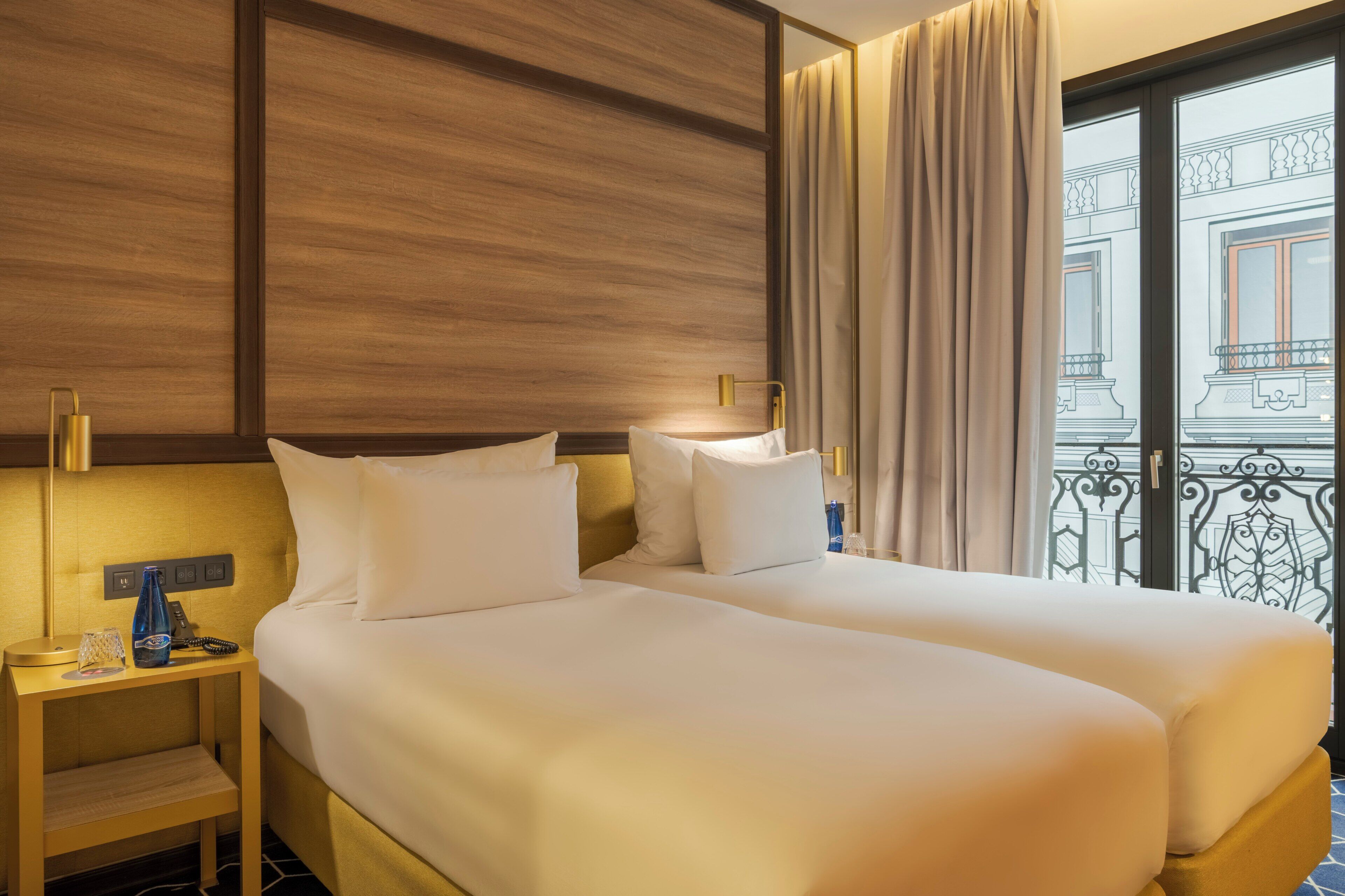 A hotel room with two beds, a nightstand with a lamp, a telephone, and a window with a view of a balcony.