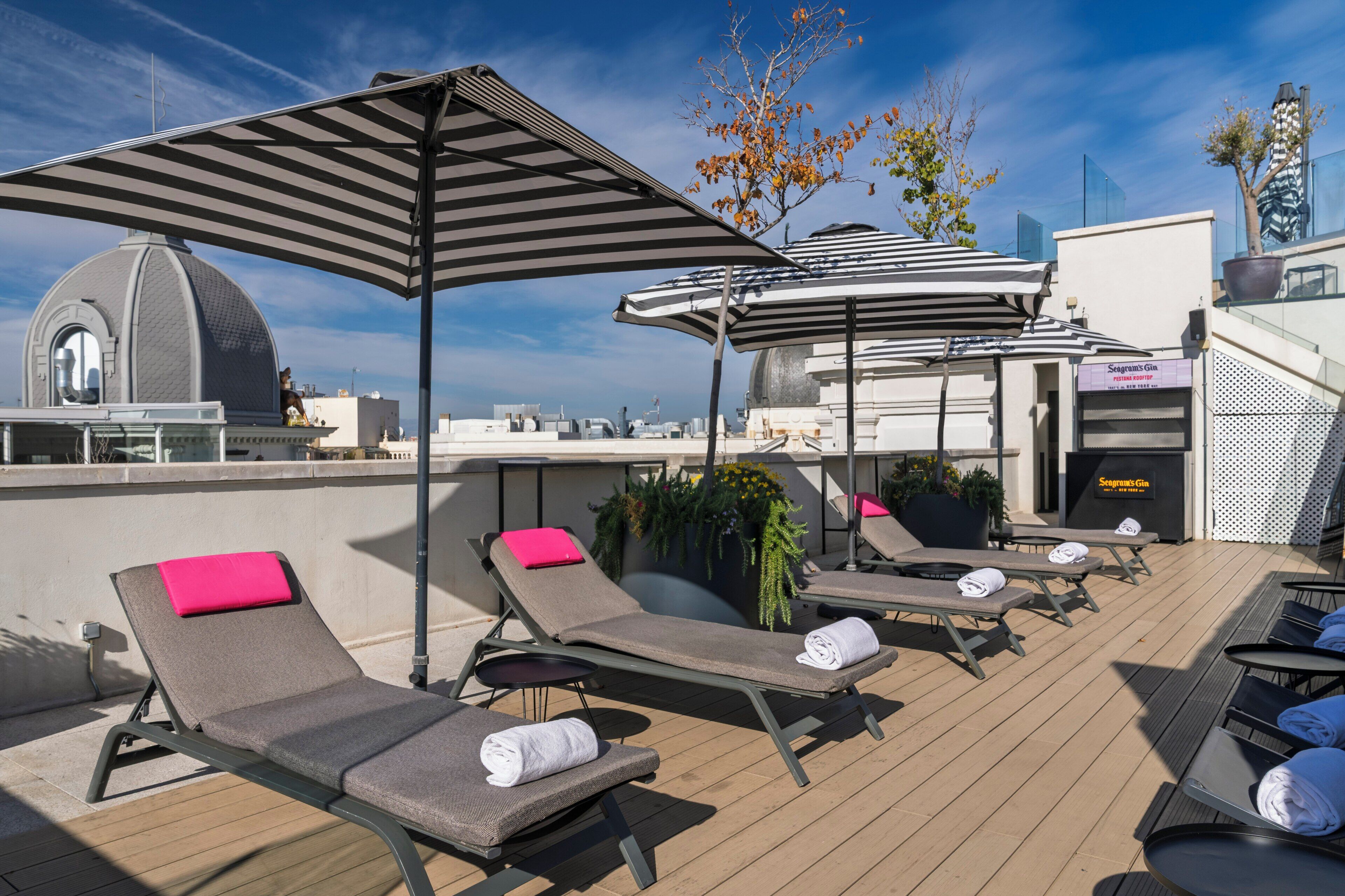 Un área de lounge en la azotea con sillones de sol, sombrillas a rayas y vistas de la ciudad.