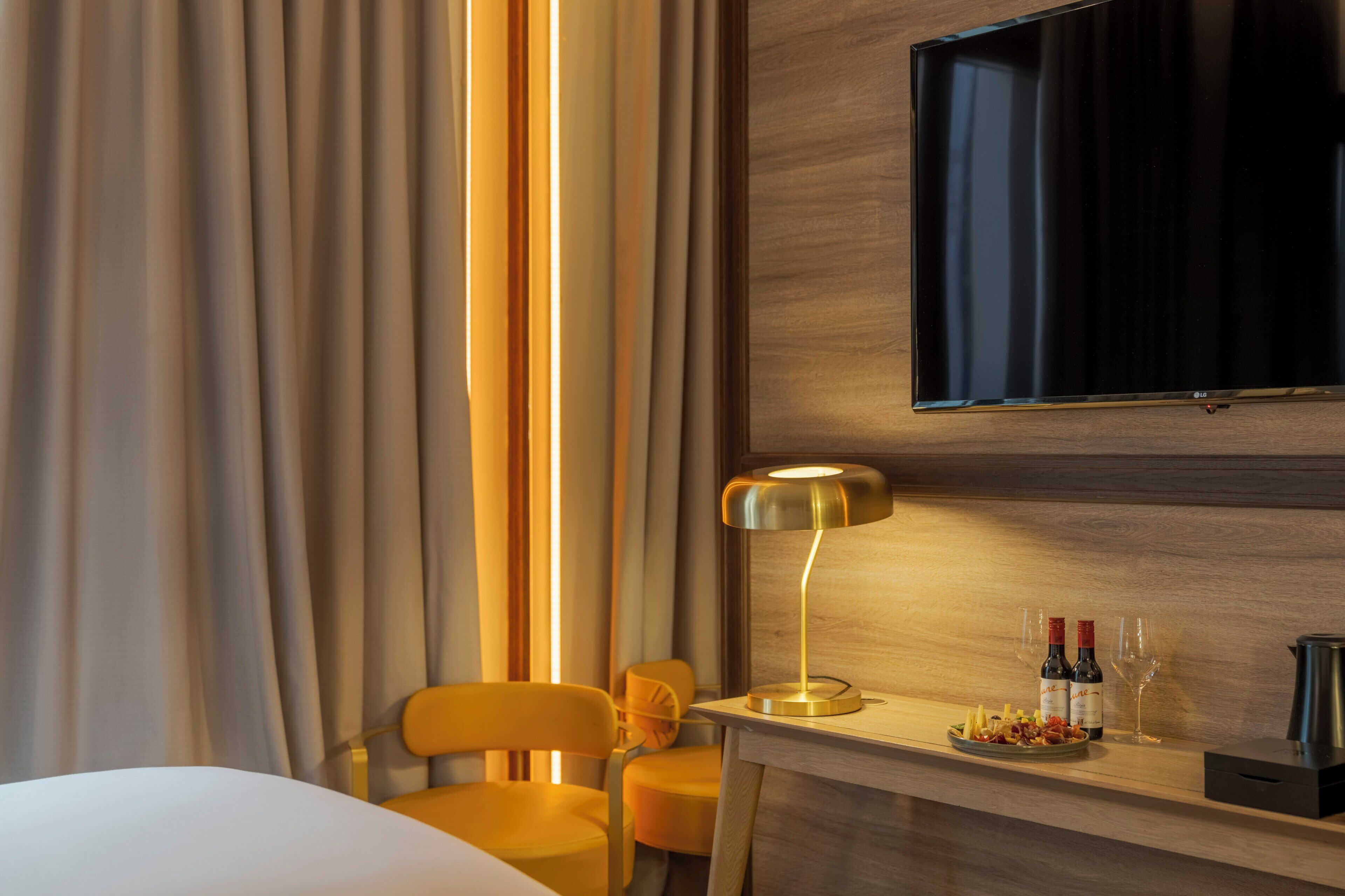 A hotel room with a bed, a desk with a lamp, a TV mounted on the wall, and a chair.
