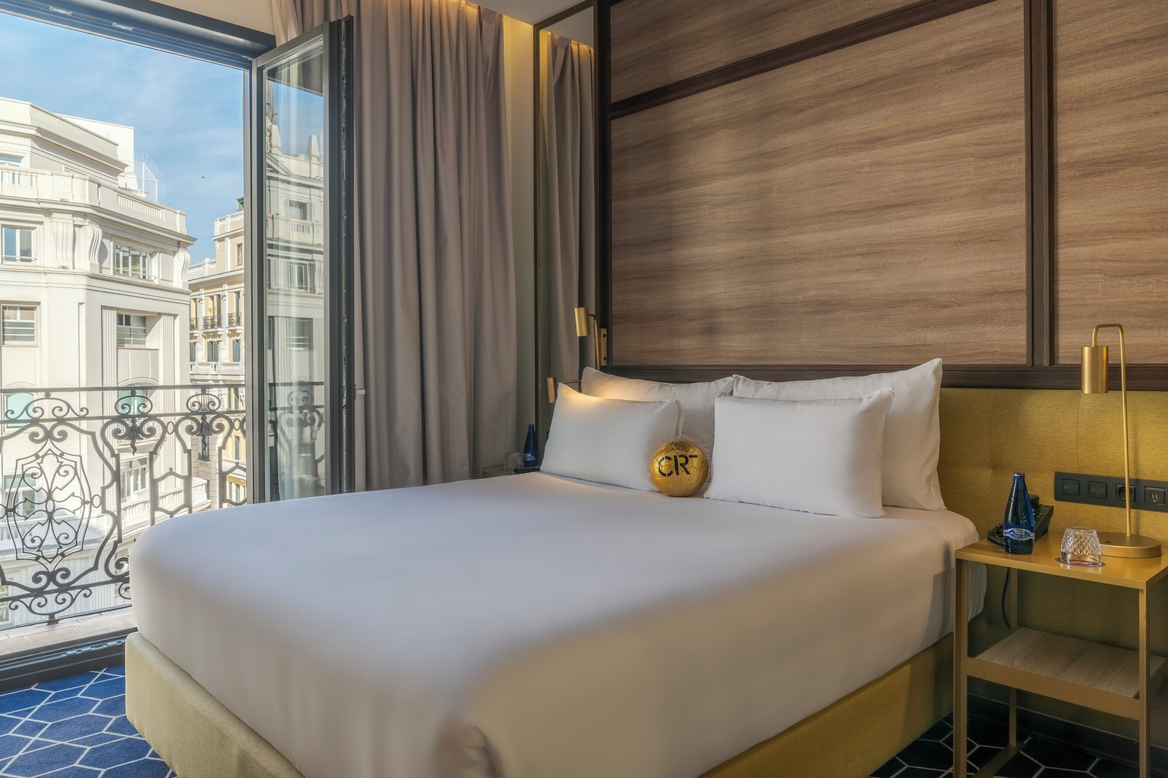 A hotel room with a large bed, a nightstand, a lamp, and a view of a cityscape through a window with curtains.