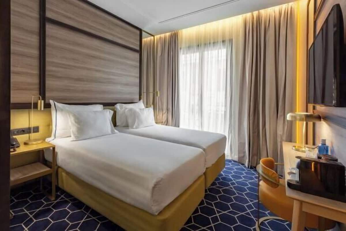 A hotel room with two beds, a desk, a TV, and a large window with curtains.