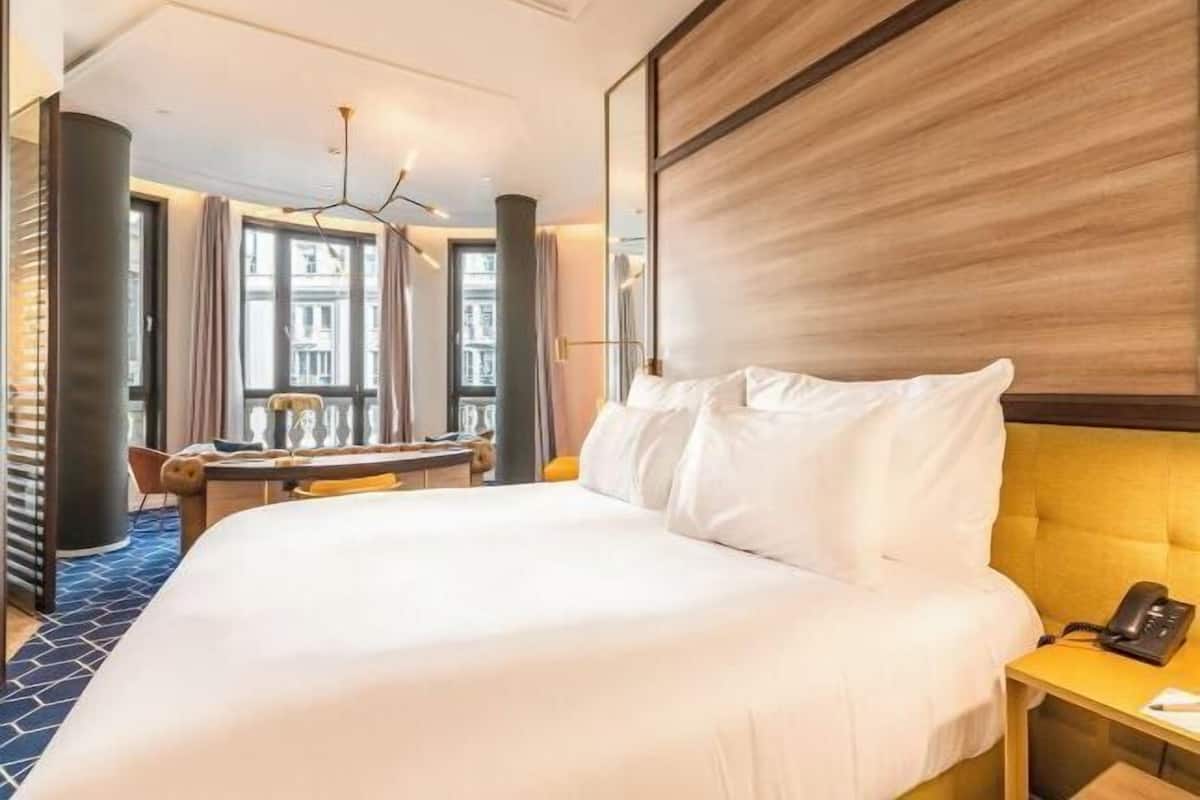 A hotel room with a large bed, a desk, a chair, and a view of buildings through the windows.