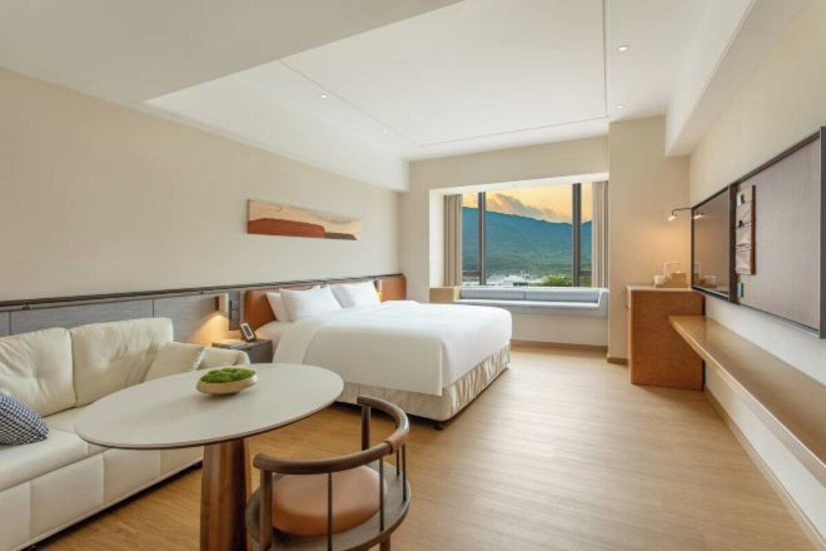 A modern hotel room with a bed, a sofa, a small table, and a large window offering a view of the mountains.
