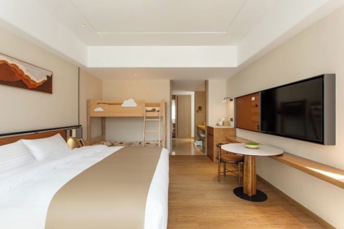 A modern hotel room with a large bed, a bunk bed, a desk with a chair, a flat-screen TV, and a painting on the wall.