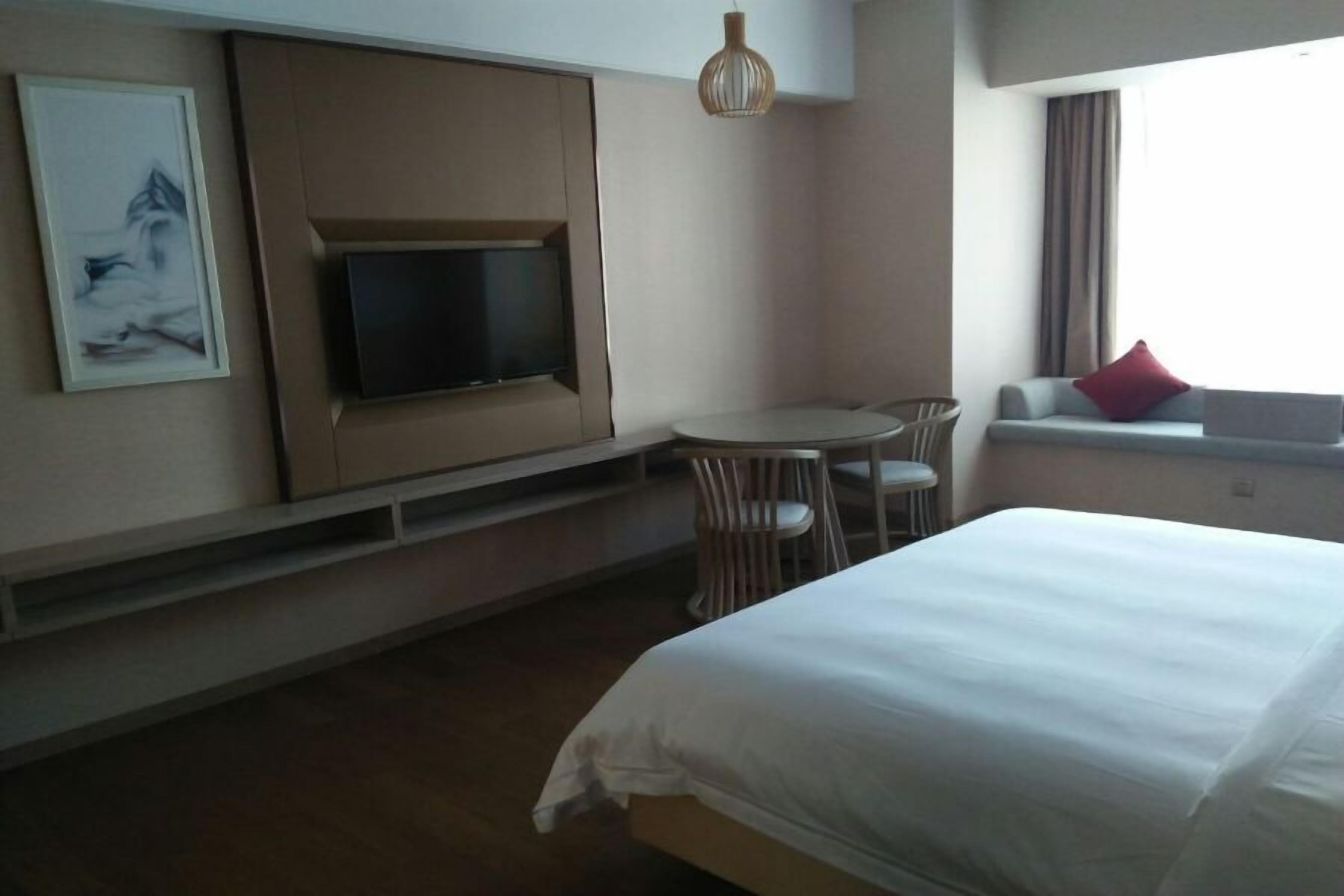 Une chambre d’hôtel avec un lit, un téléviseur fixé au mur, une petite table avec des chaises et une fenêtre agrémentée d’un coussin rouge.