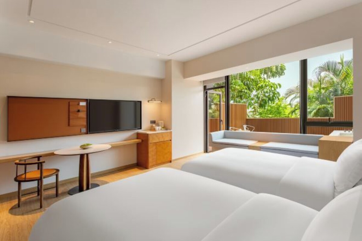 A modern hotel room with a large bed, a desk with a chair, a TV, and a window with a view of greenery.