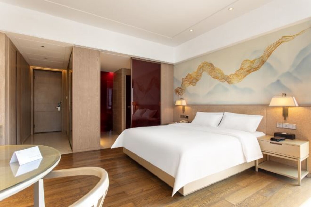 A hotel room with a large bed, bedside tables, a desk, and a chair. There is a painting on the wall and a door leading to another room.