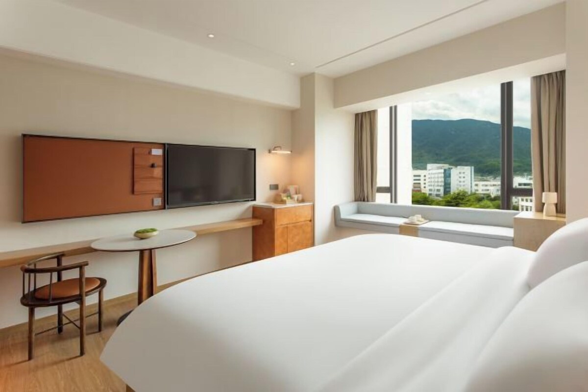 Une chambre d’hôtel avec un grand lit, un bureau avec une chaise, une télévision et une fenêtre donnant sur des immeubles et des montagnes.
