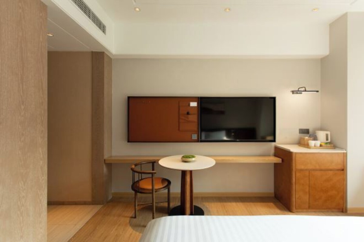 A modern hotel room with a bed, a small round table with a chair, a TV mounted on the wall, and a wooden cabinet.