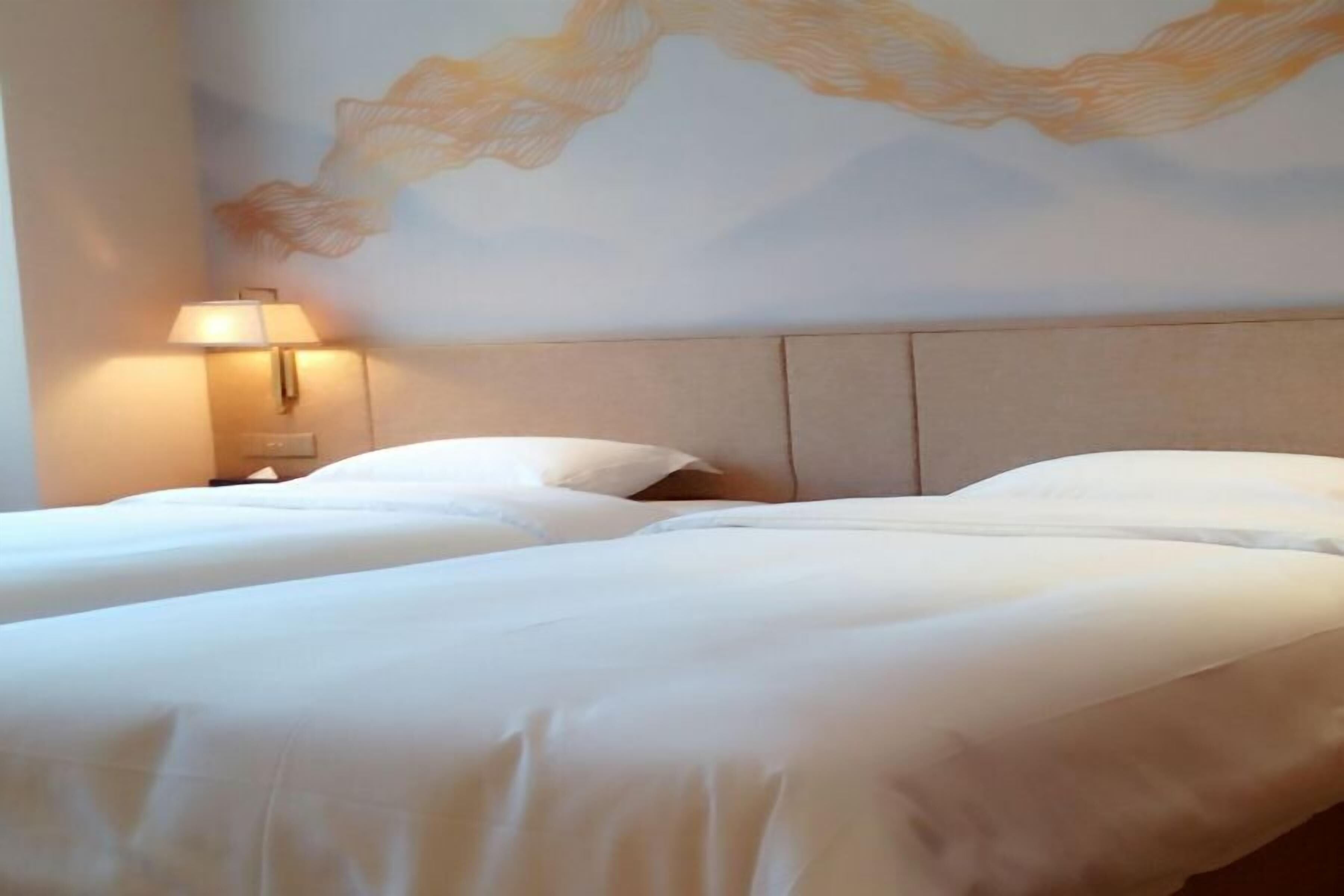 Quarto de hotel com duas camas, uma mesa de cabeceira, um abajur e um mural na parede.