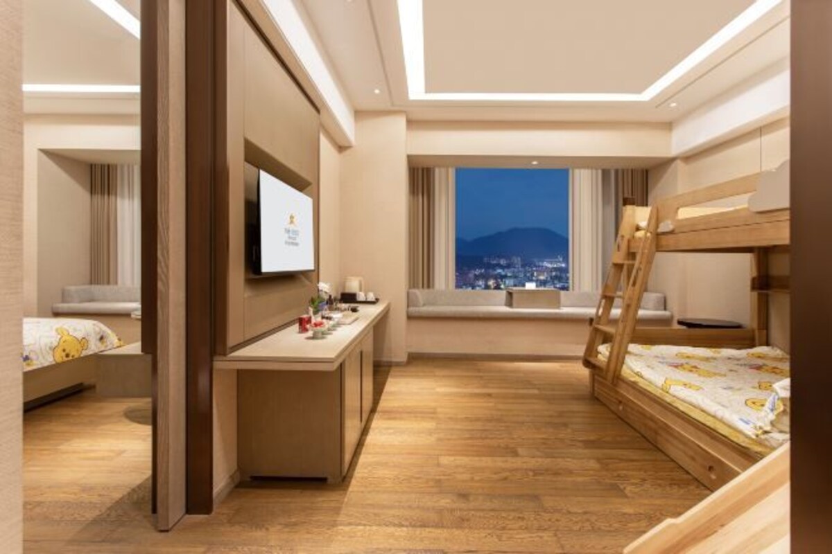 A modern hotel room with a wooden bunk bed, a desk with a flat-screen TV, and a large window offering a city view.