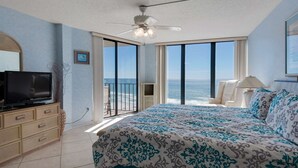 A bedroom with a large bed, a TV on a wooden dresser, and a balcony with a view of the ocean.