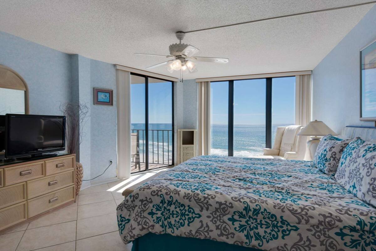 Ein Schlafzimmer mit einem großen Bett, einem Fernseher auf einer Holzkommode und einem Balkon mit Meerblick.