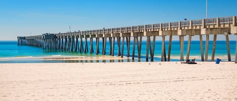 A pier extending into the ocean with a flag at the end, a sandy beach, and a clear sky.