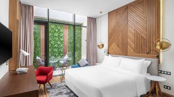 Habitación de hotel moderna con una cama grande, un escritorio, una silla roja y vistas a zonas verdes a través de grandes ventanales.