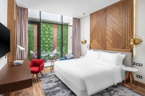A modern hotel room with a large bed, a desk, a red chair, and a view of greenery through large windows.