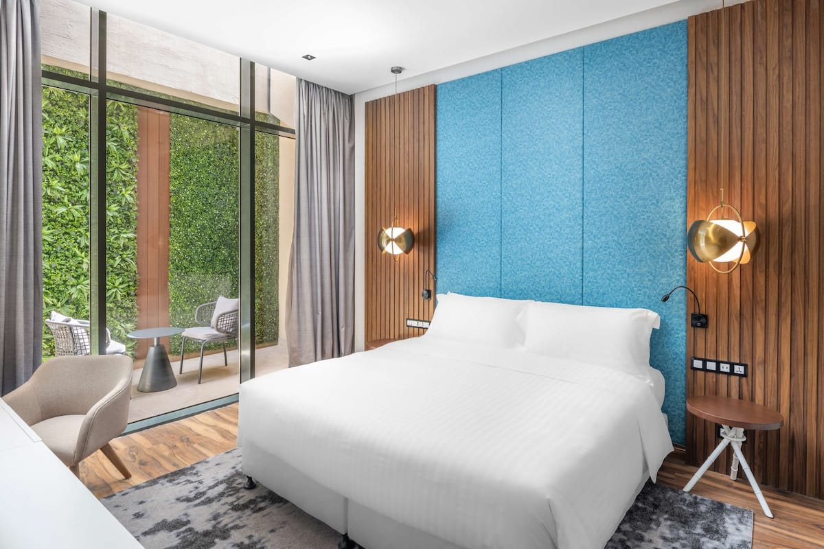 A modern hotel room with a large bed, a small table, a chair, and a view of greenery through large windows.