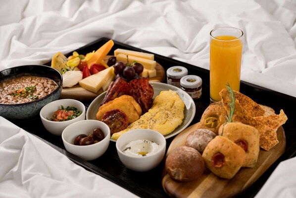 A breakfast tray with various foods and a glass of orange juice on a bed with white sheets.