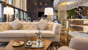 Un hall d'hôtel moderne avec un canapé confortable, une table basse sur laquelle est disposée une sélection de pâtisseries, et un bar en arrière-plan.