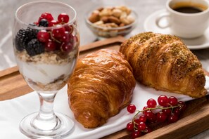 A glass of yogurt parfait with berries, two croissants, and a cup of coffee on a wooden board.