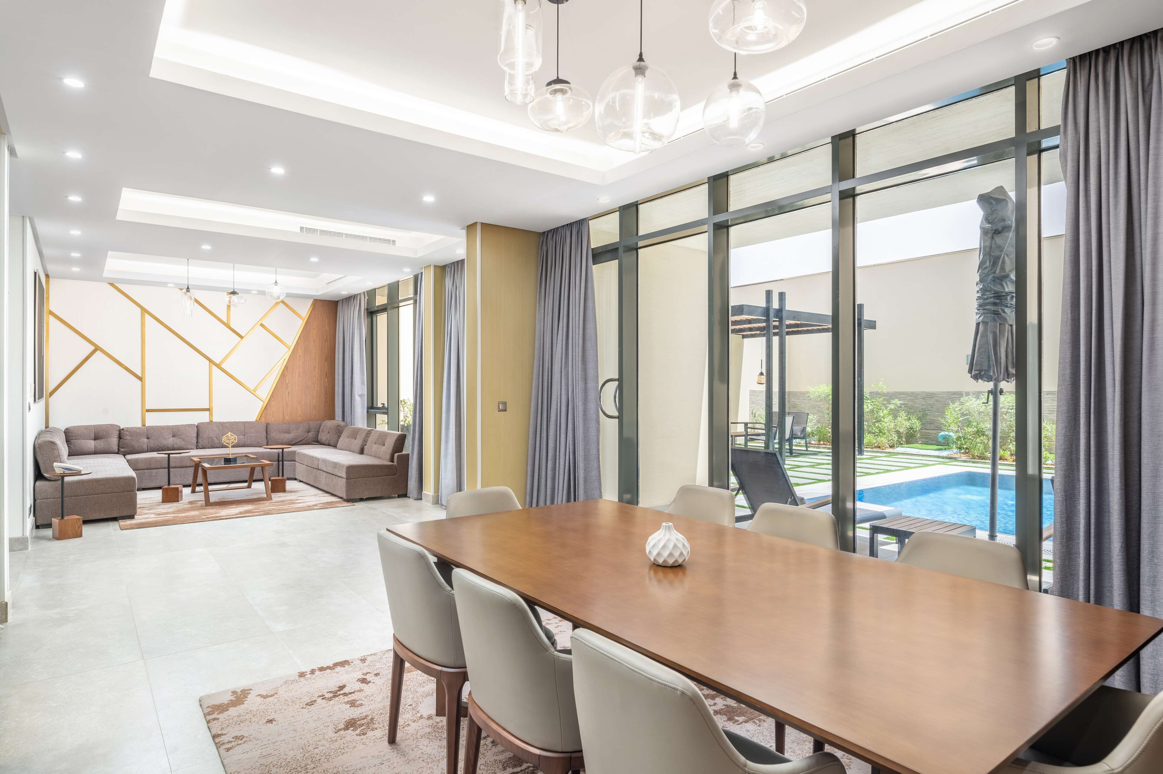 A modern living room with a large sectional sofa, a wooden dining table with chairs, and a view of a swimming pool through large windows.