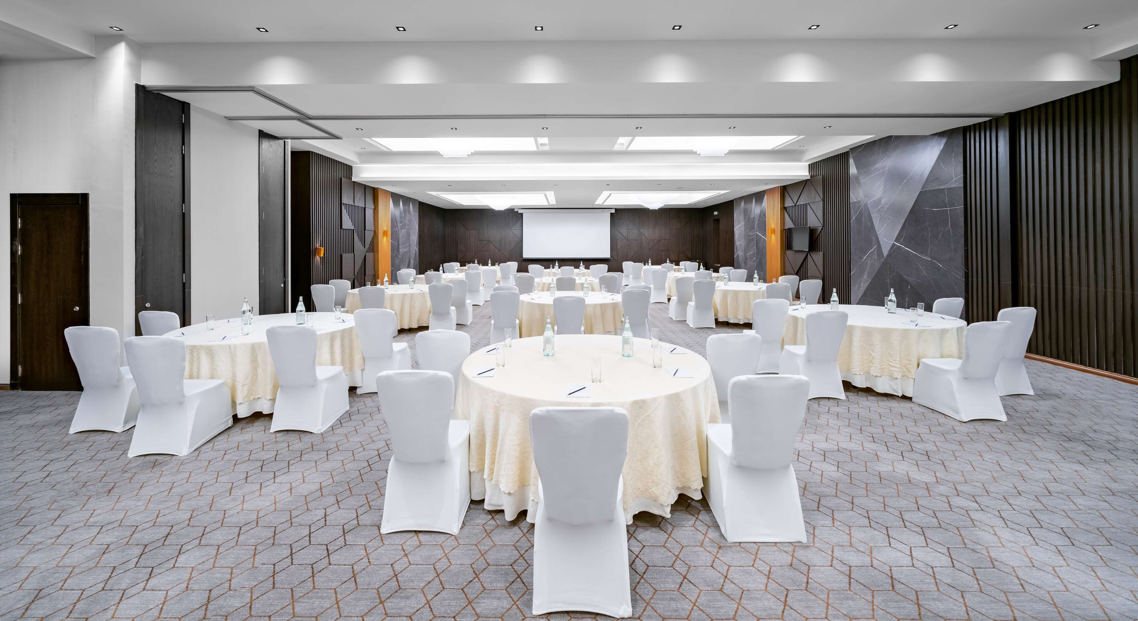 A conference room with multiple round tables, white chairs, and a large screen at the front.