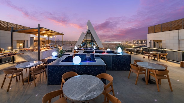 Un bar situé sur un toit, doté d’un design moderne, avec un espace salon central comprenant des tables et des chaises rondes, un comptoir de bar et une vue sur les toits de la ville au crépuscule.