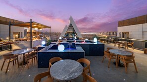 A rooftop bar with a modern design, featuring a central seating area with round tables and chairs, a bar counter, and a view of the city skyline at dusk.