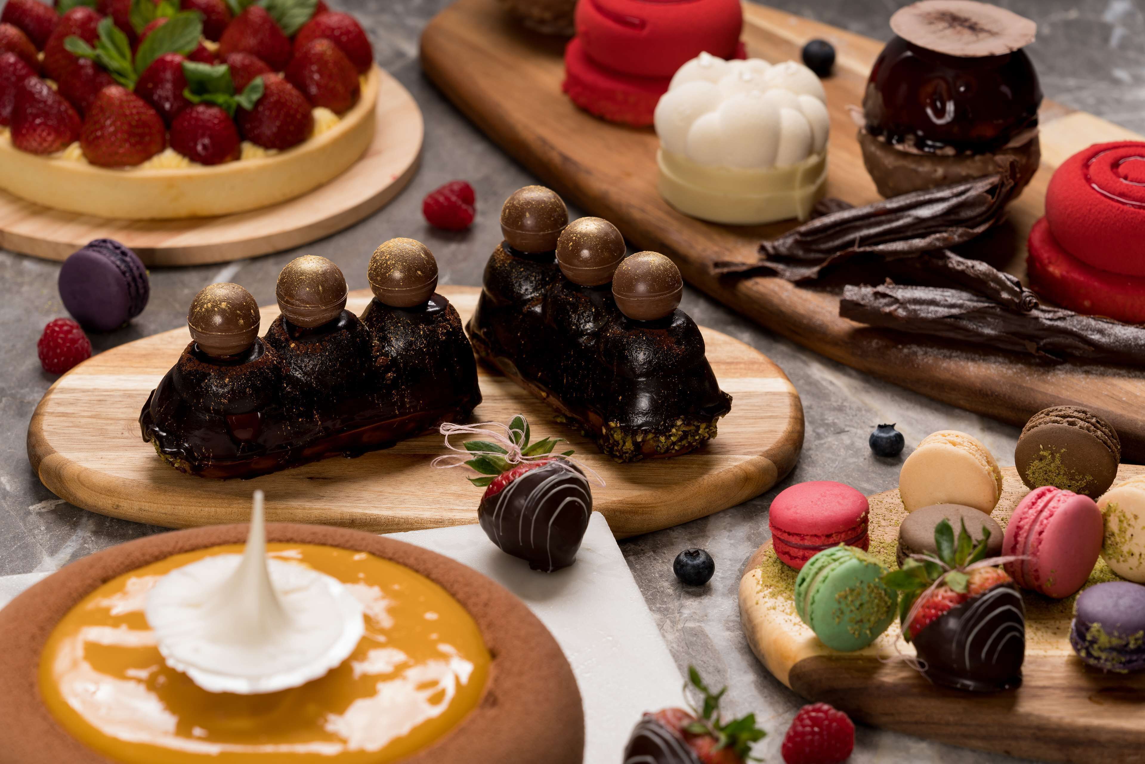 Variedad de postres en tablas de madera, entre ellos fresas cubiertas de chocolate, macarons y una tarta de fresas.