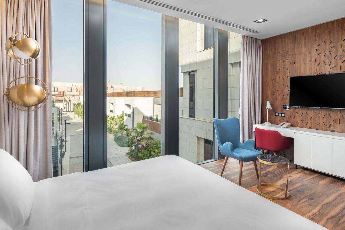 A modern hotel room with a large bed, a desk with a chair, a TV, and a view of the city through large windows.