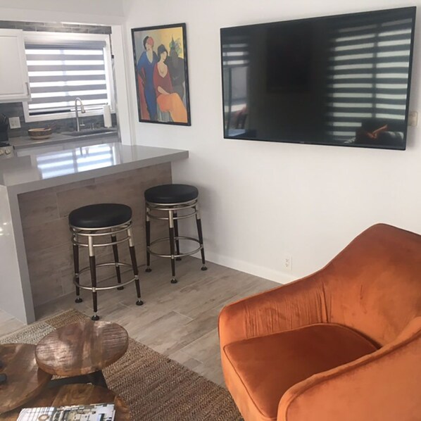 A modern kitchen with a bar area, two stools, a large flat-screen TV, and an orange chair. - location, location, location (Long Beach)