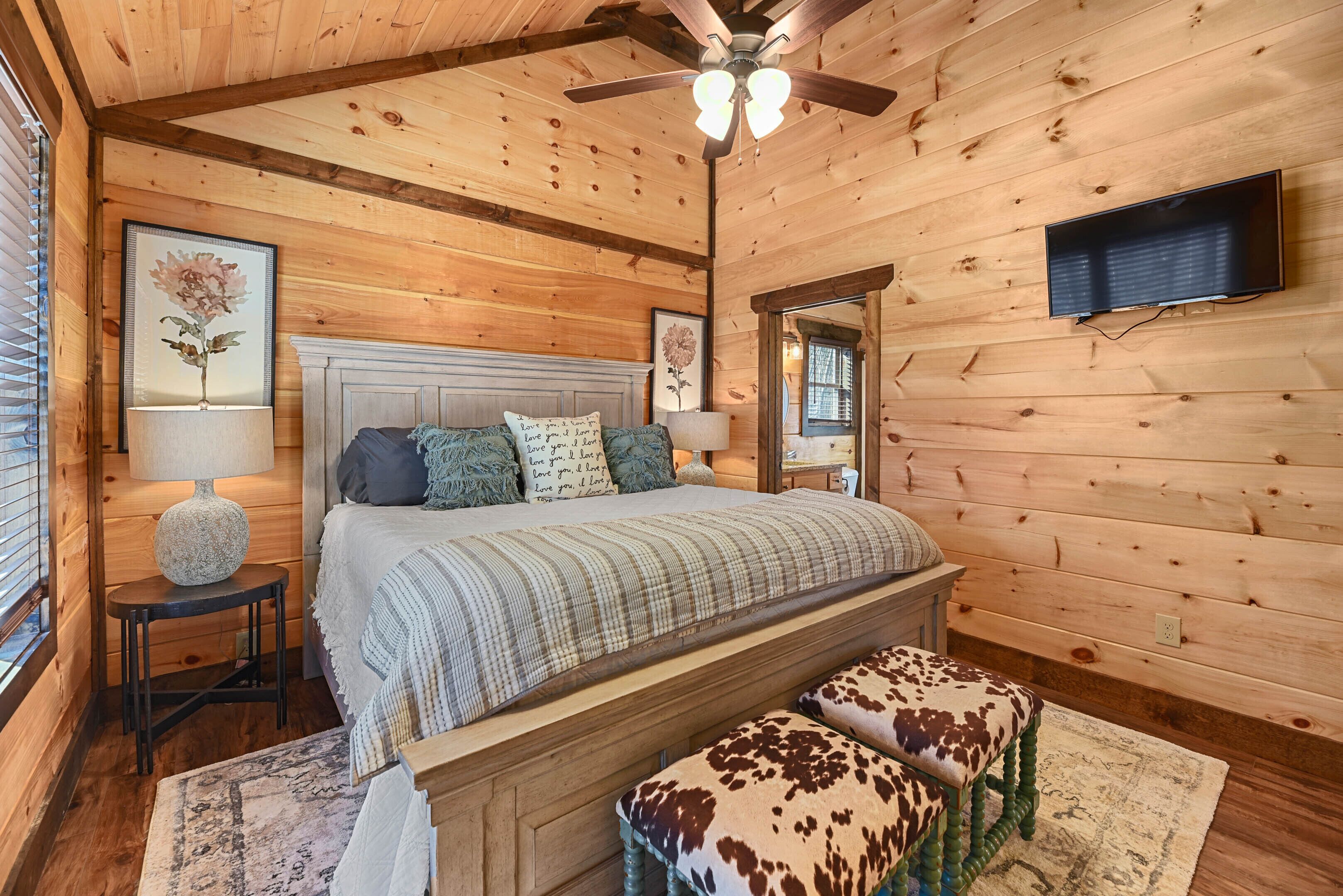 A wooden cabin bedroom with a bed, two nightstands, a ceiling fan, and a mounted TV.