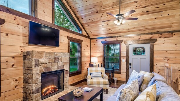 L’intérieur chaleureux d’un chalet avec une cheminée en pierre, des murs en bois, un ventilateur de plafond et un téléviseur fixé au mur.
