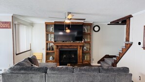 Una sala de estar con un sofá seccional gris, un mueble de entretenimiento de madera con un televisor de pantalla plana, una chimenea y una escalera de madera con un rascador para gatos.