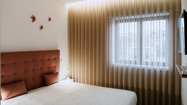 Habitación de hotel con cabecera capitonada, cama con ropa de cama blanca y ventana con persianas verticales.