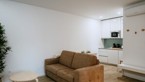 Una sala de estar moderna con un sofá marrón, una mesita redonda y una zona de cocina con armarios blancos y un microondas.