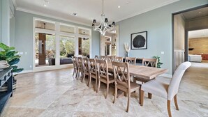 A spacious dining area with a large wooden table and chairs, a chandelier, and large windows offering a view of the outdoors. - Luxurious Secluded Estate with Pool & Pond (Galveston)