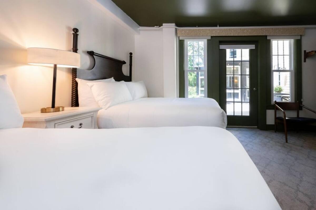 A hotel room with two beds, a nightstand with a lamp, a chair, and a view of the outside through a door.