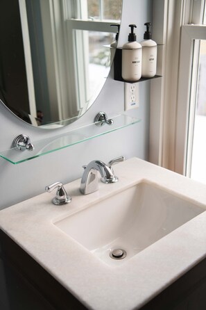 A bathroom with a white sink, two faucets, a glass shelf above the sink, and a mirror reflecting the window.