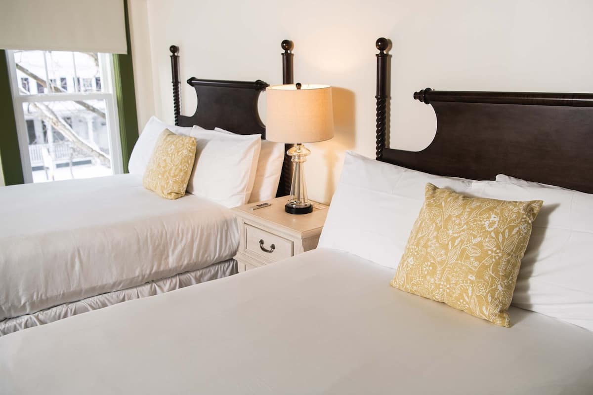 A hotel room with two beds, a nightstand, a lamp, and a window with a view of buildings.
