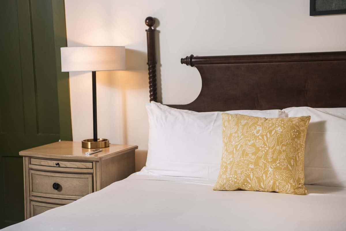 A neatly made bed with white and patterned pillows, a wooden nightstand with a lamp, and a green door in the background.