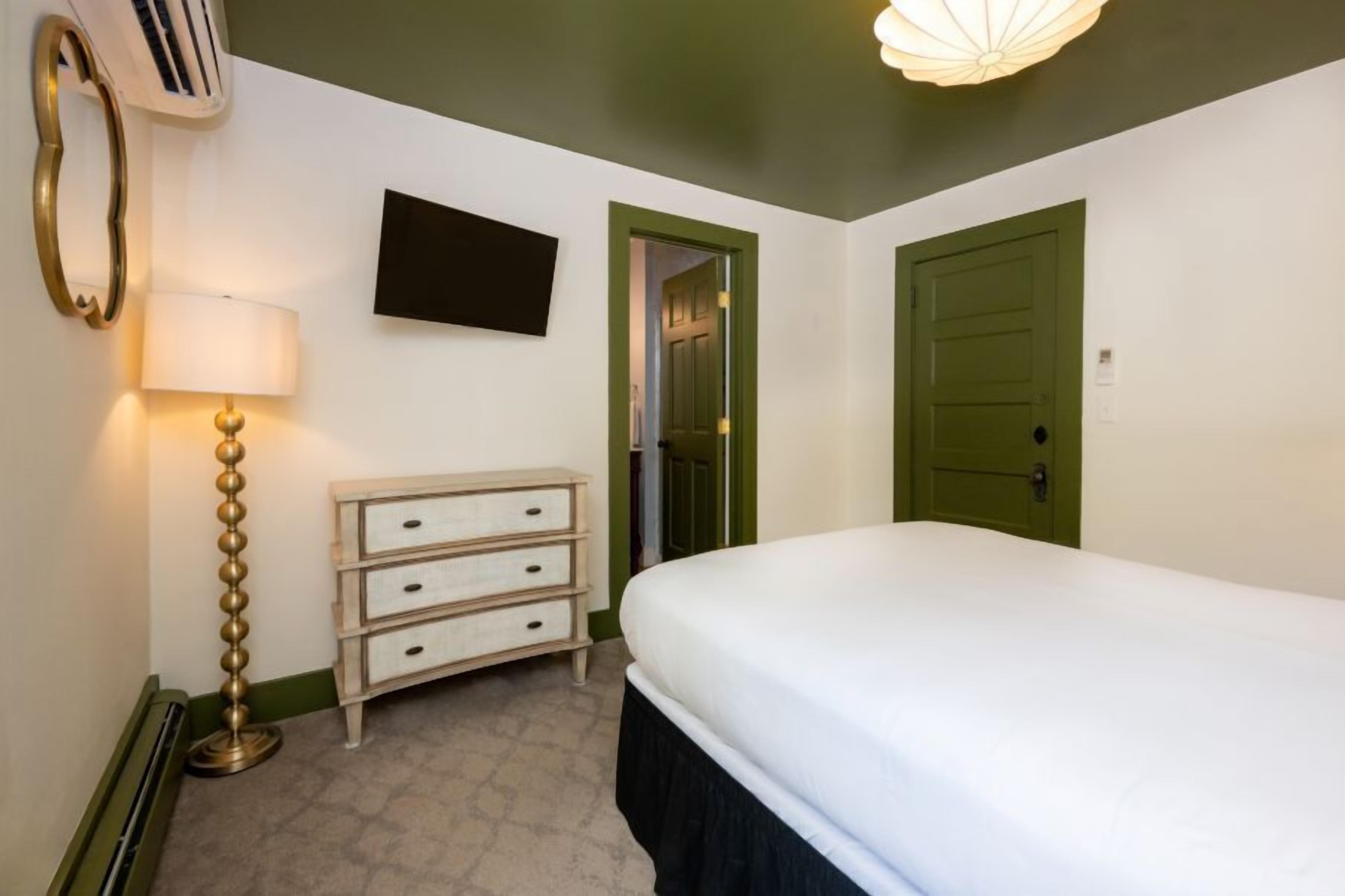 A bedroom with a bed, a dresser, a lamp, a TV, and a door.