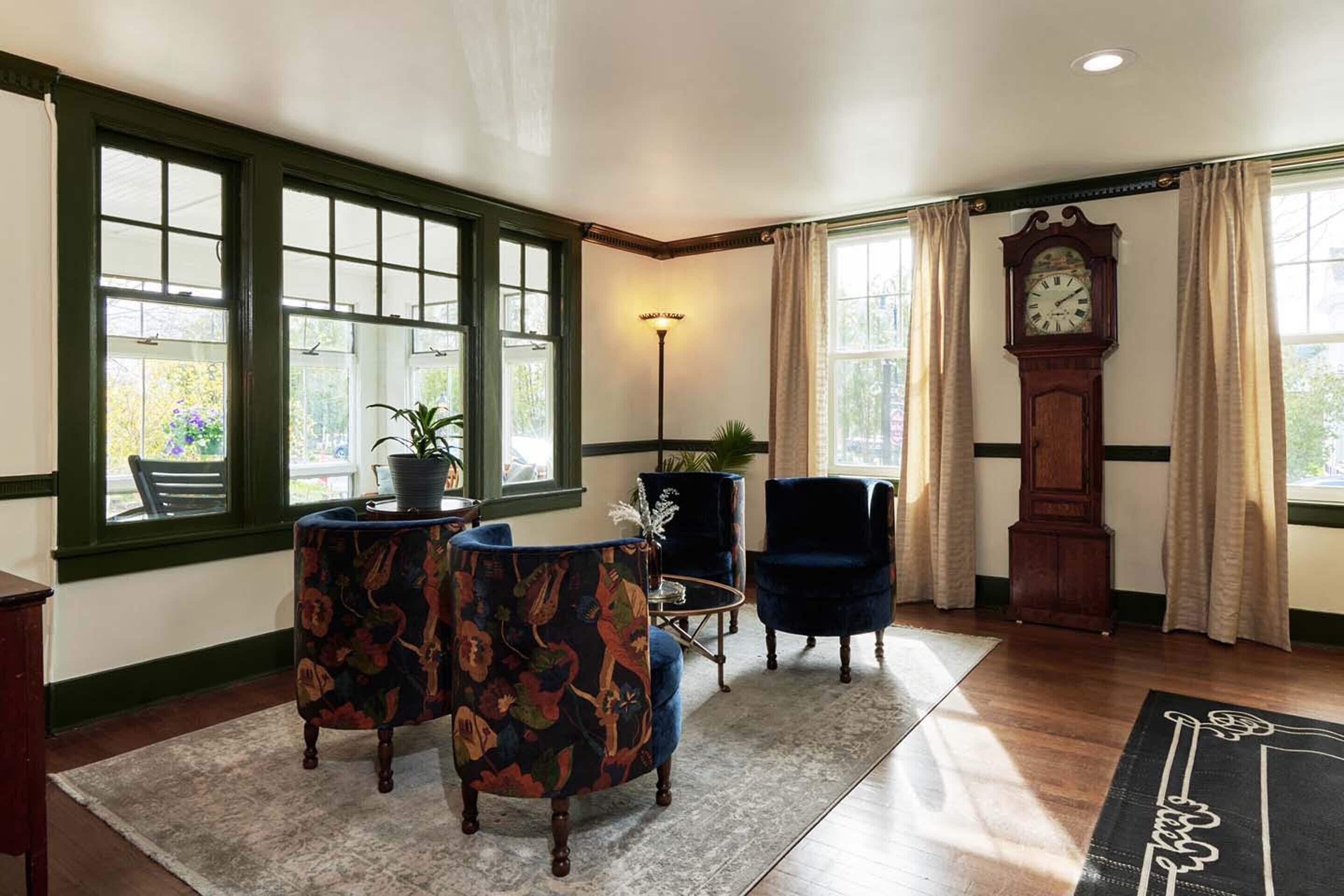 A room with a large window, a clock, a blue armchair, a patterned armchair, and a small table with a plant.