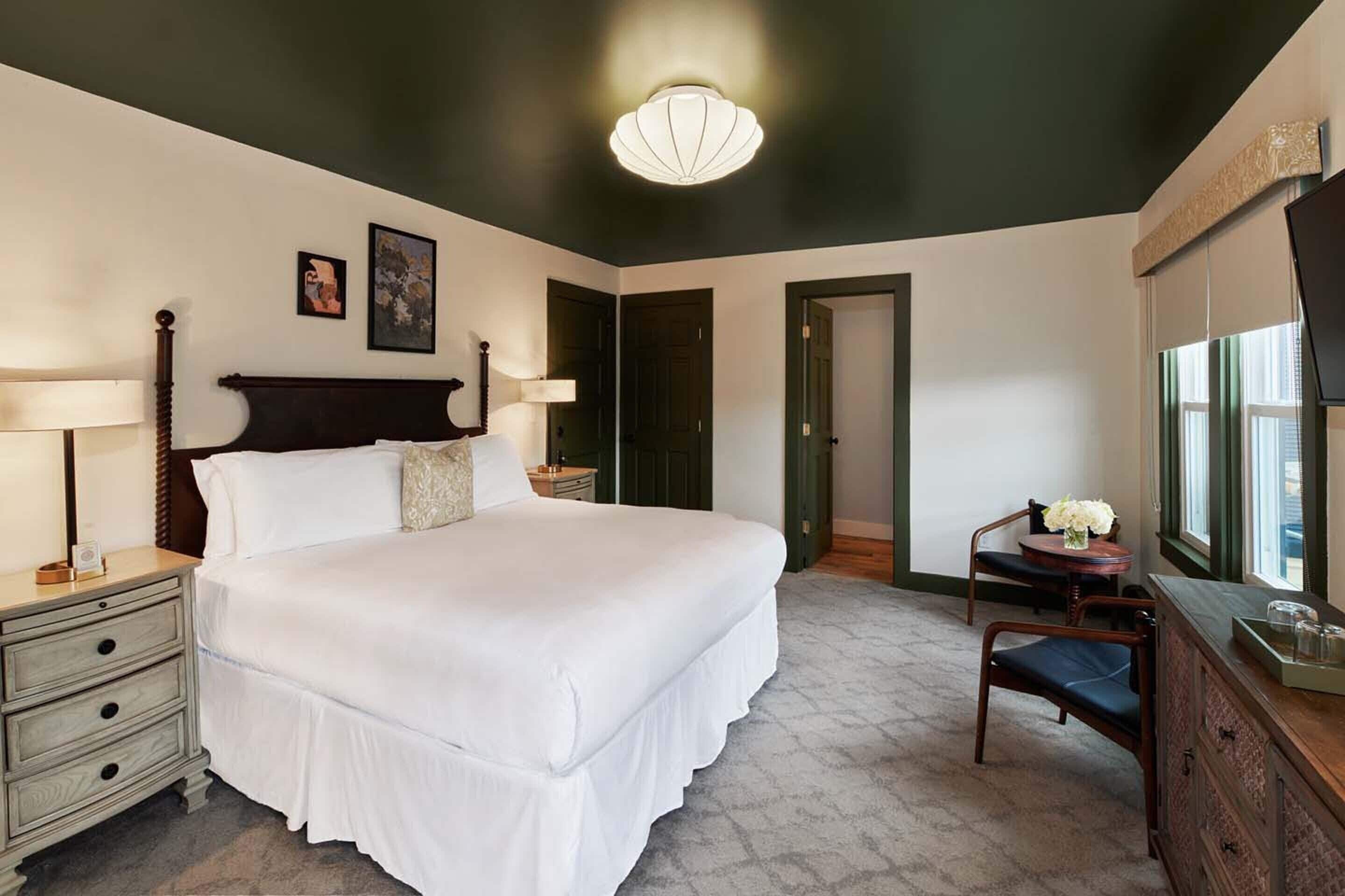 Un dormitorio con una cama grande, dos mesitas de noche con lámparas, un aparador, una silla y un escritorio con un jarrón de flores.