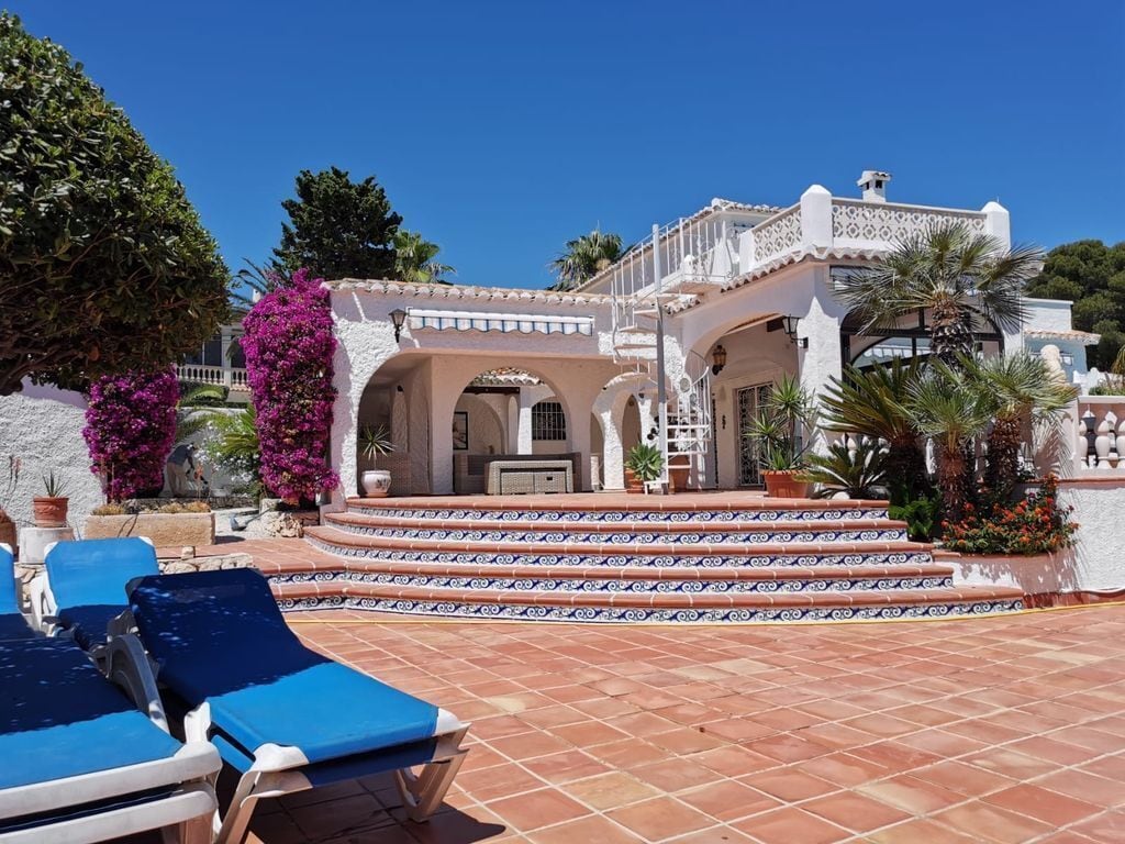 A villa with a tiled patio, blue lounge chairs, and a white building with arches and a balcony.